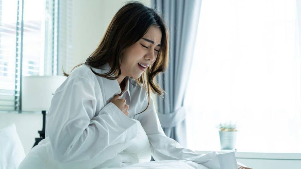 A woman in a white shirt sits on a bed, clutching her chest with a pained expression.