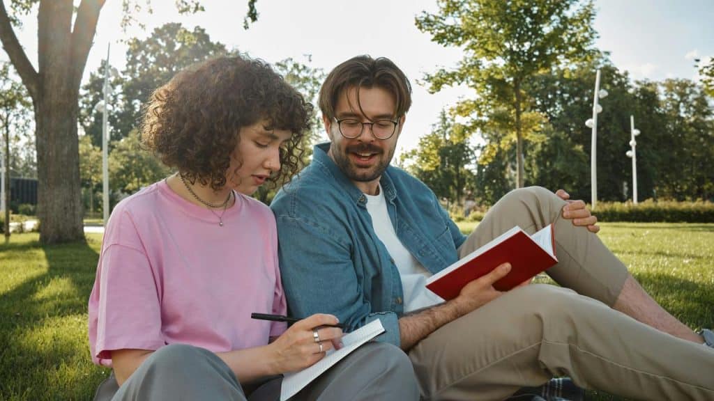 A couple sitting on grass in a park, reading and writing together.