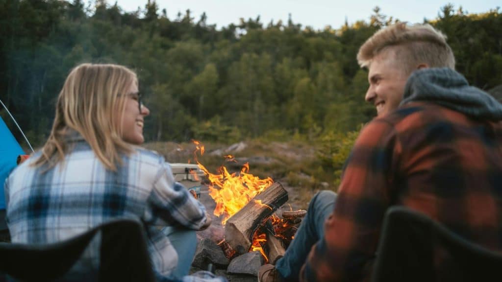 A couple sitting by a campfire outdoors, smiling and talking in a forest setting.