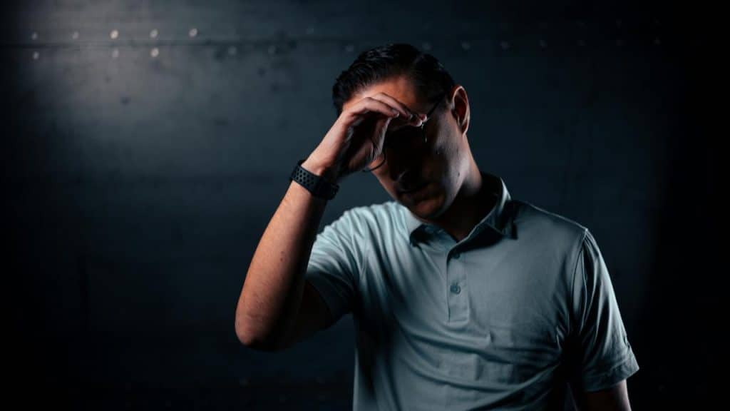 A man standing against a dark background with one hand on his forehead.