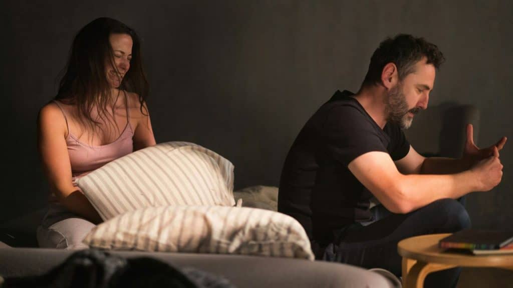 A man and woman sit on a bed in a dimly lit room, facing away from each other.
