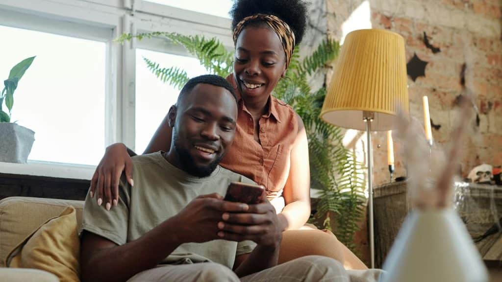 A couple smiling while looking at a phone together in a bright, plant-filled living room.