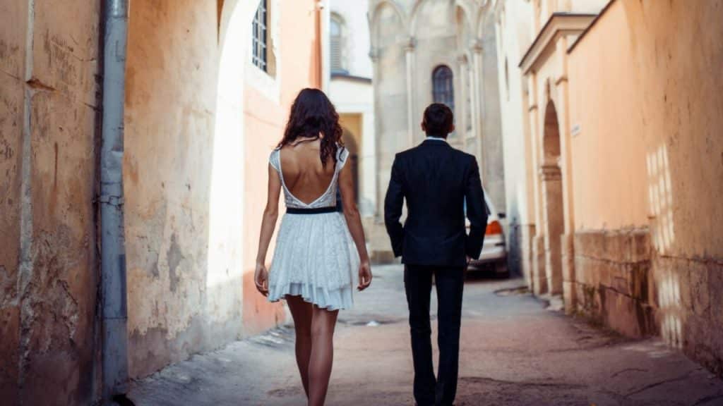 A woman in a white dress and a man in a suit walk down a narrow alleyway.