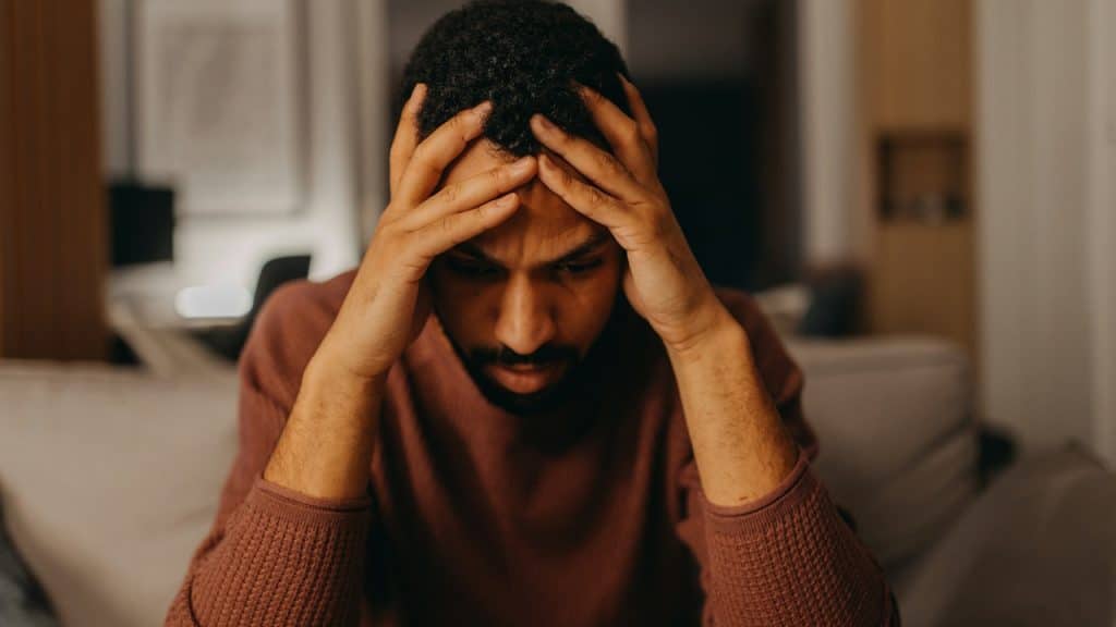 A bearded man sitting on a couch looks worried and holds his head in his hands.