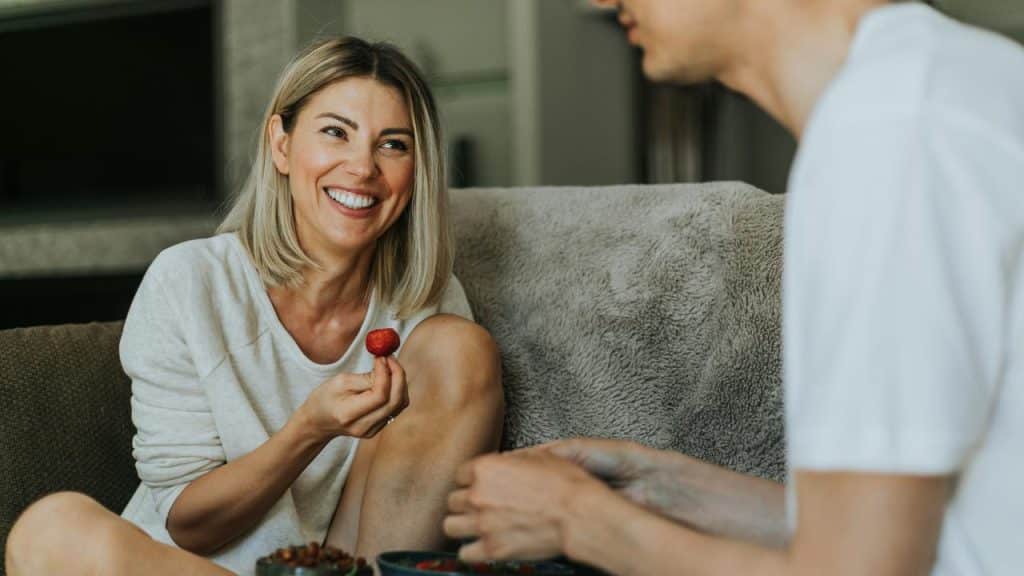 A woman smiling on a couch while holding a strawberry.