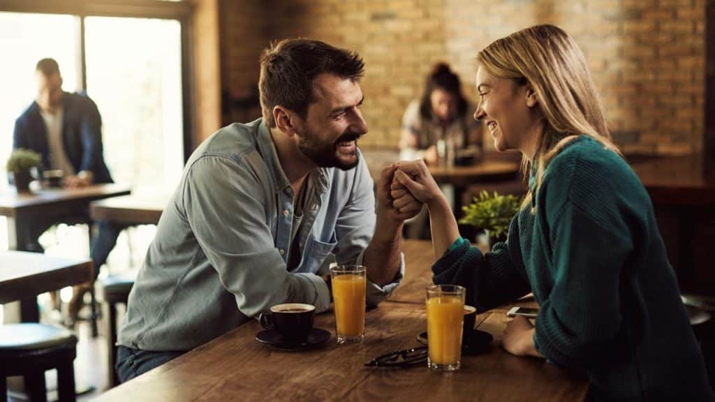 A man and woman sit at a cafe table holding hands and looking at each other.