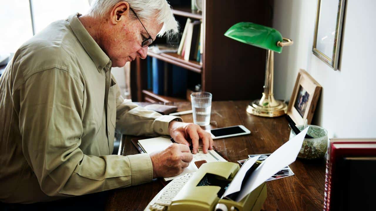An elderly man writing at a desk with a typewriter.