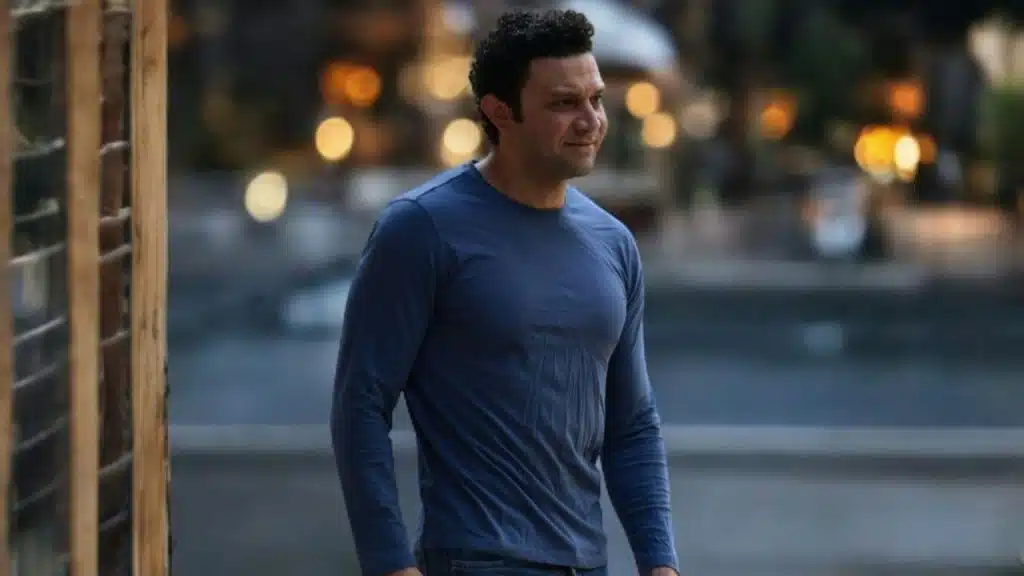 A man in a long-sleeved blue shirt looks toward the side in a blurry outdoor setting.