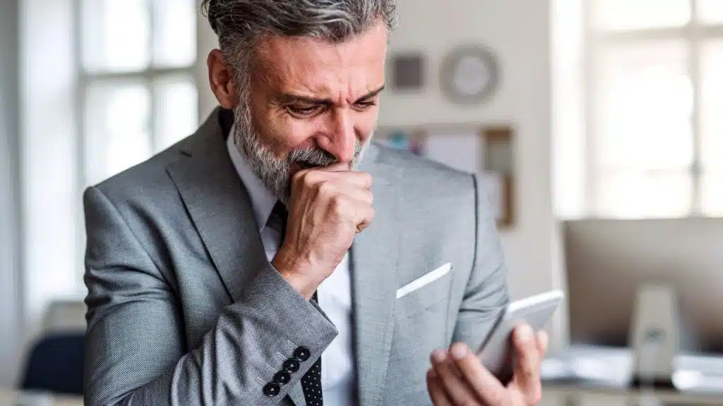 A grey-haired man in a suit looks at a smartphone while biting his knuckles anxiously.