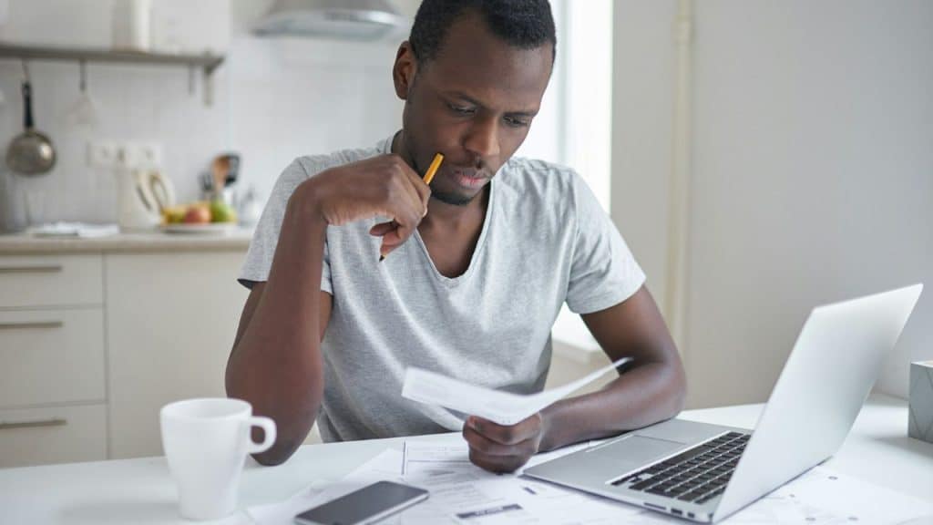 A man in a gray shirt sits at a white desk with a laptop, looking at a paper.
