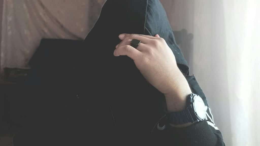 A person in dark clothing resting a hand with a ring and watch near their face.
