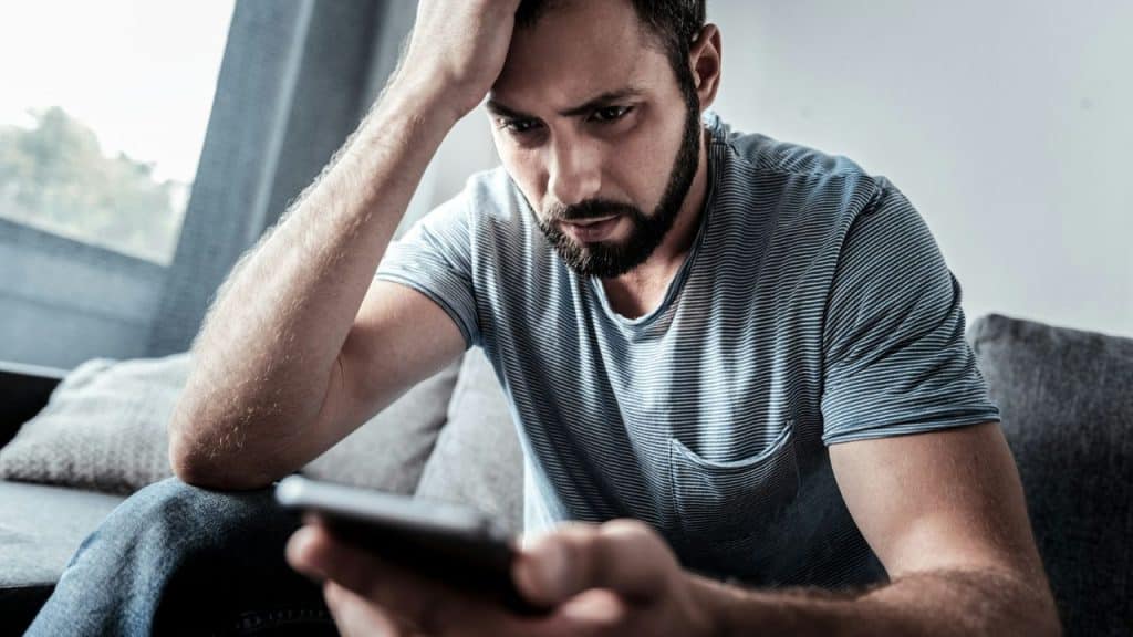 A bearded man sits on a couch holding his head while looking down at a smartphone.