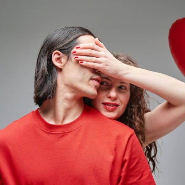 A woman with red nails covers a man's eyes; two red heart balloons float nearby.