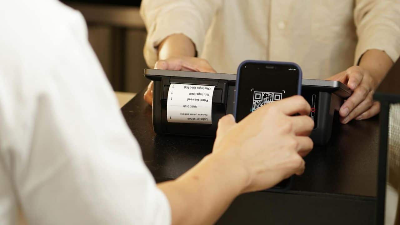 A person uses a smartphone to scan a QR code on a digital payment terminal screen.
