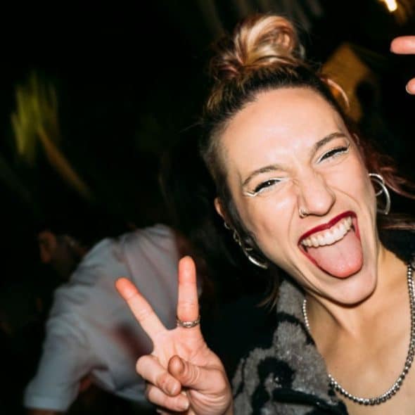 A smiling person sticks their tongue out while making peace signs in a dark setting.