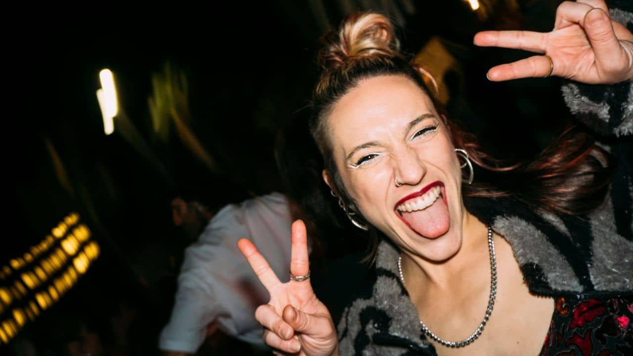 A smiling person sticks their tongue out while making peace signs in a dark setting.