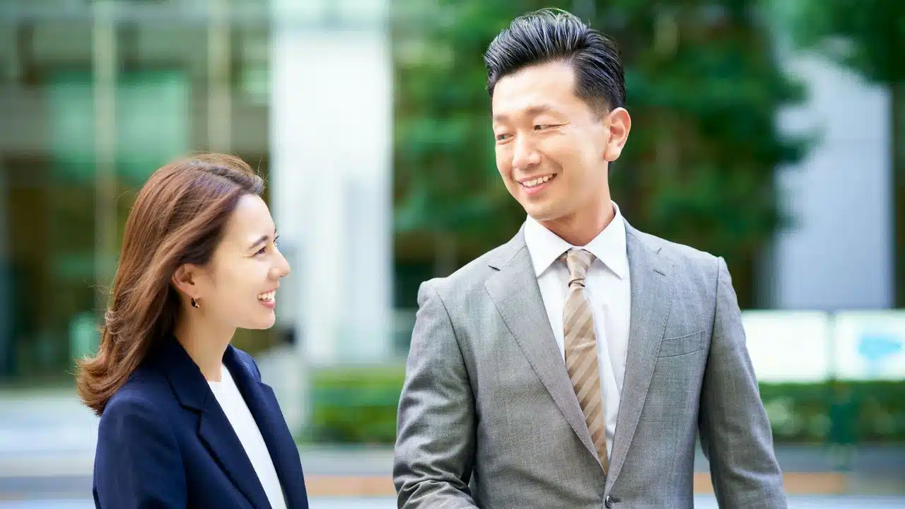 A man and a woman in business attire smile while looking at each other outdoors.