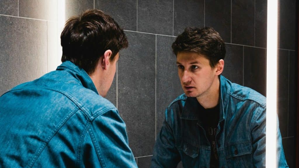 A man in a denim jacket stares at his own reflection in a bathroom mirror.