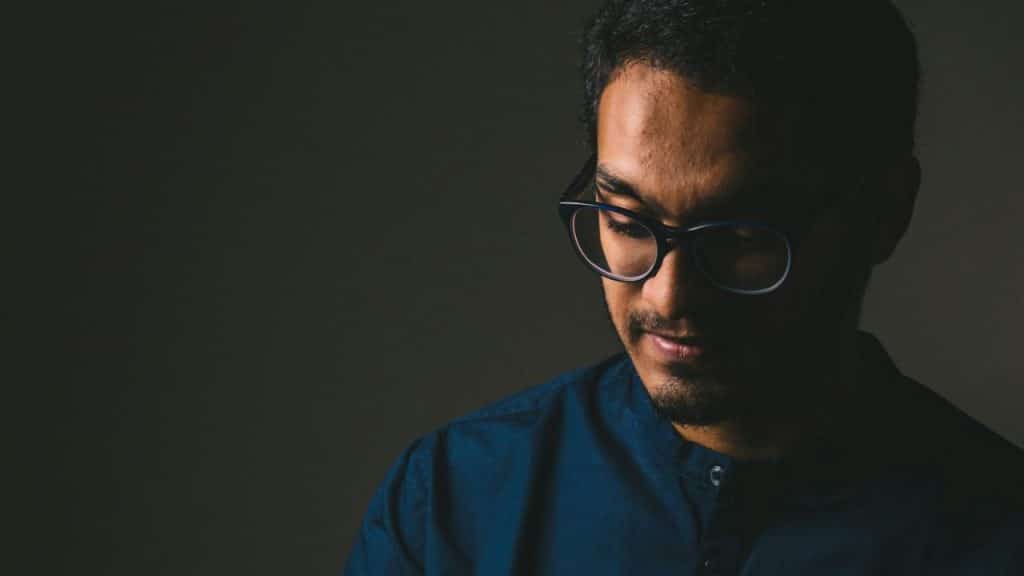 A man with glasses and a goatee looks downward against a dark, solid background.