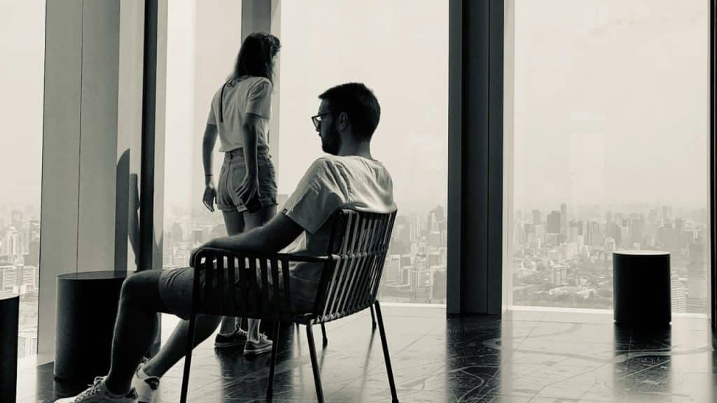 A man sits in a chair indoors while a woman stands nearby looking out a window.