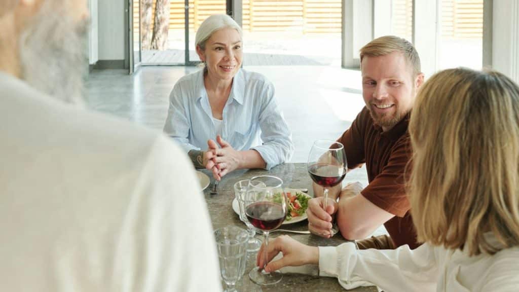 Several people sit around a table with food and wine, looking toward an unseen person.