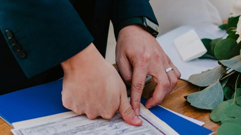 A close-up of hands signing a document on a table with greenery nearby.