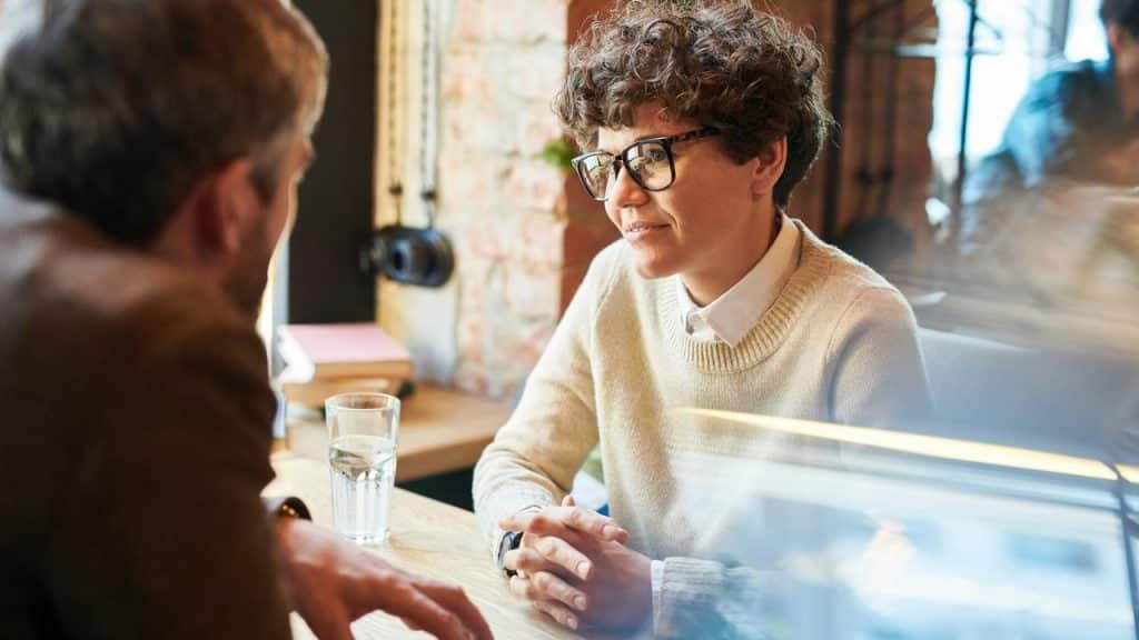A person in glasses listening to someone while sitting at a café table.