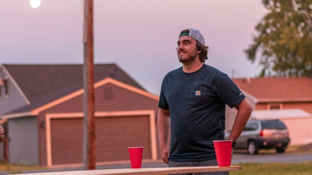 A man in a baseball cap stands outdoors at dusk near a table with two red cups.