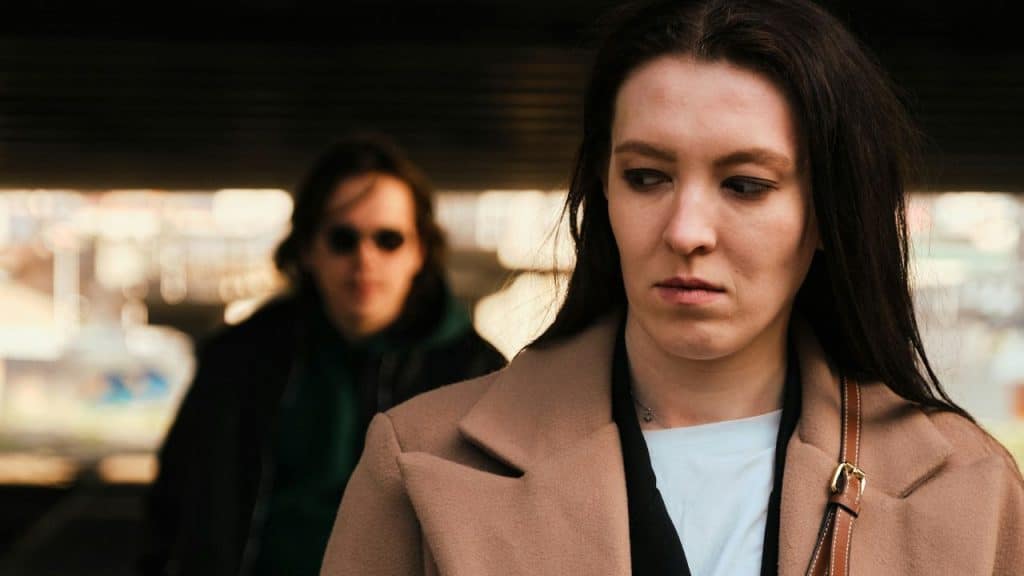 A woman in a tan coat looks away while a man stands in the background.