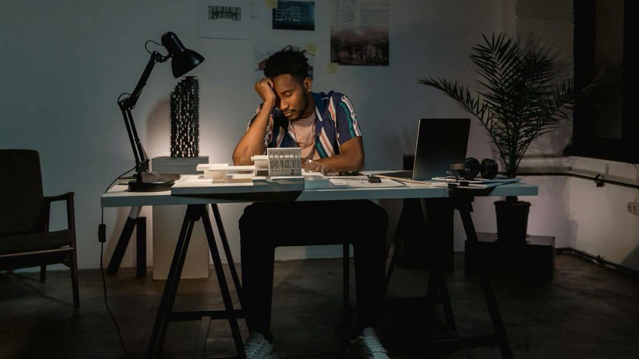 A man sits at a desk with architectural models, resting his head in his hand.