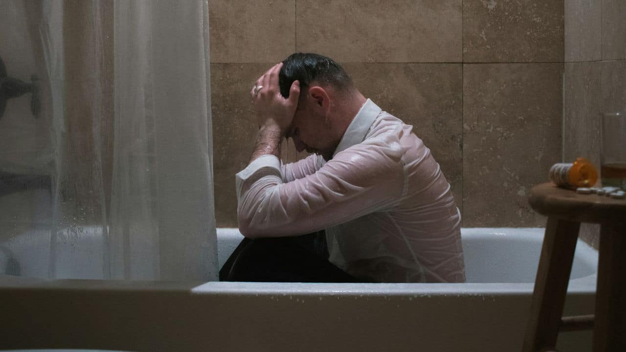 A man in a white dress shirt sits in a bathtub holding his head.
