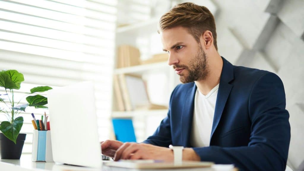 A man in a blue blazer sits at a desk while typing on a white laptop.