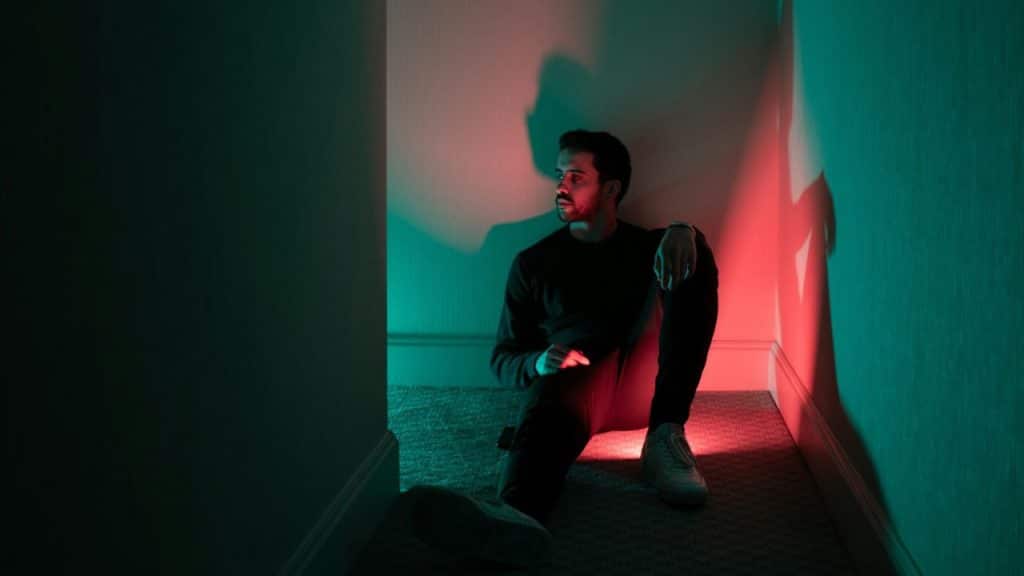 A man sits on the floor in a corner illuminated by red and green light.