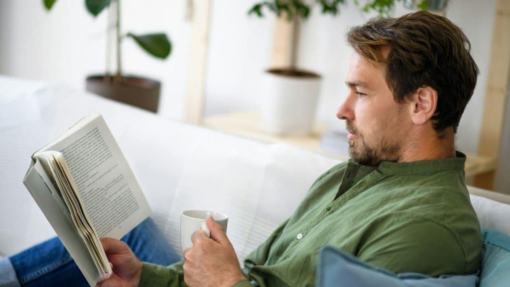 A man in a green shirt holds a white mug while reading a book indoors.