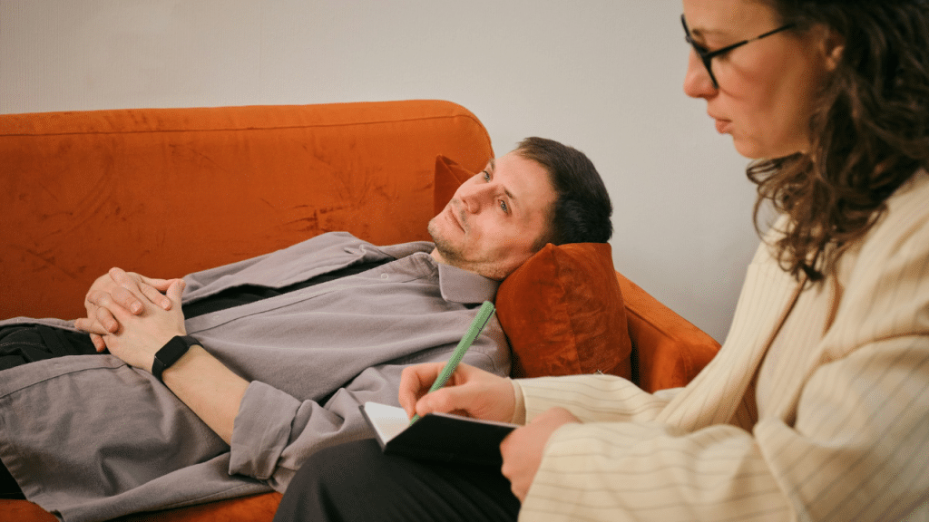 A man lies on an orange sofa while a woman takes notes in a small notebook.