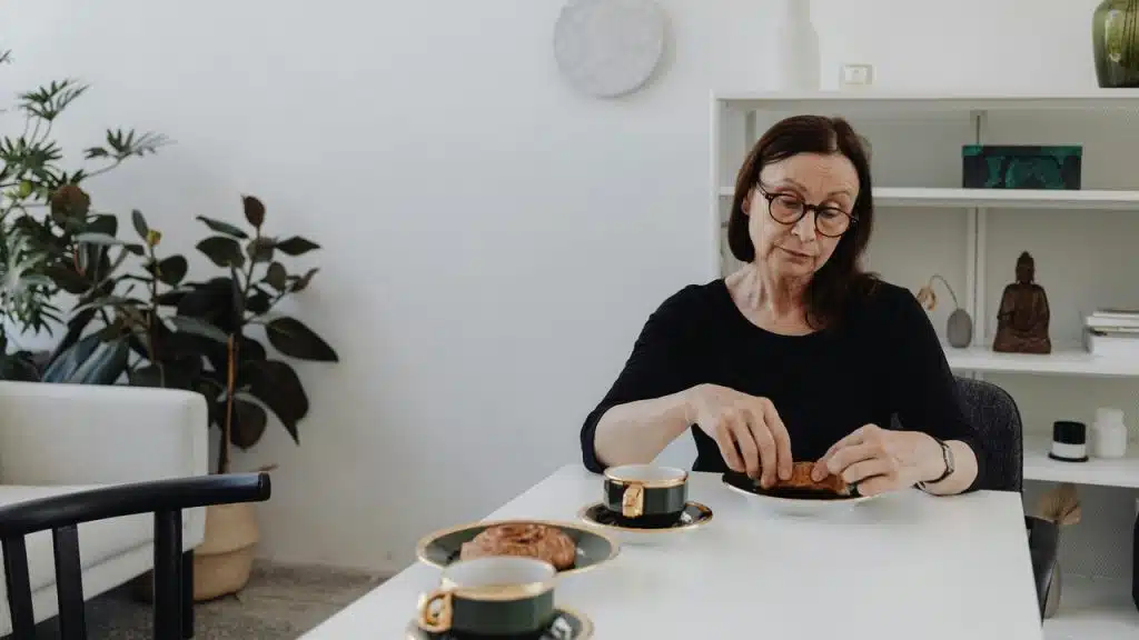 A woman sitting at a table, eating a pastry with a cup of coffee in a minimalist room.