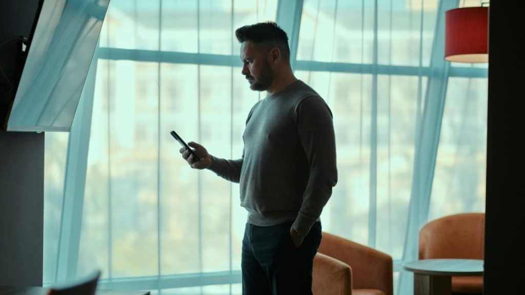 A man in a grey sweater stands indoors by a large window, looking at his smartphone.