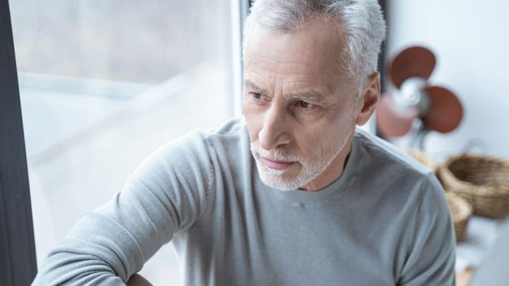 An older man with short grey hair looks out a window with a serious expression.