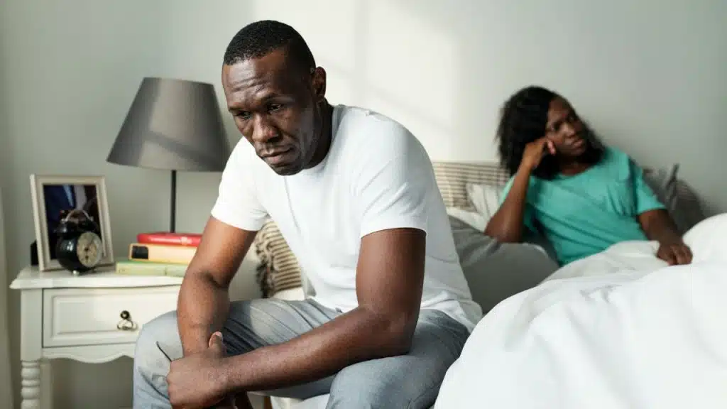 A man sits on the edge of a bed while a woman watches from behind.