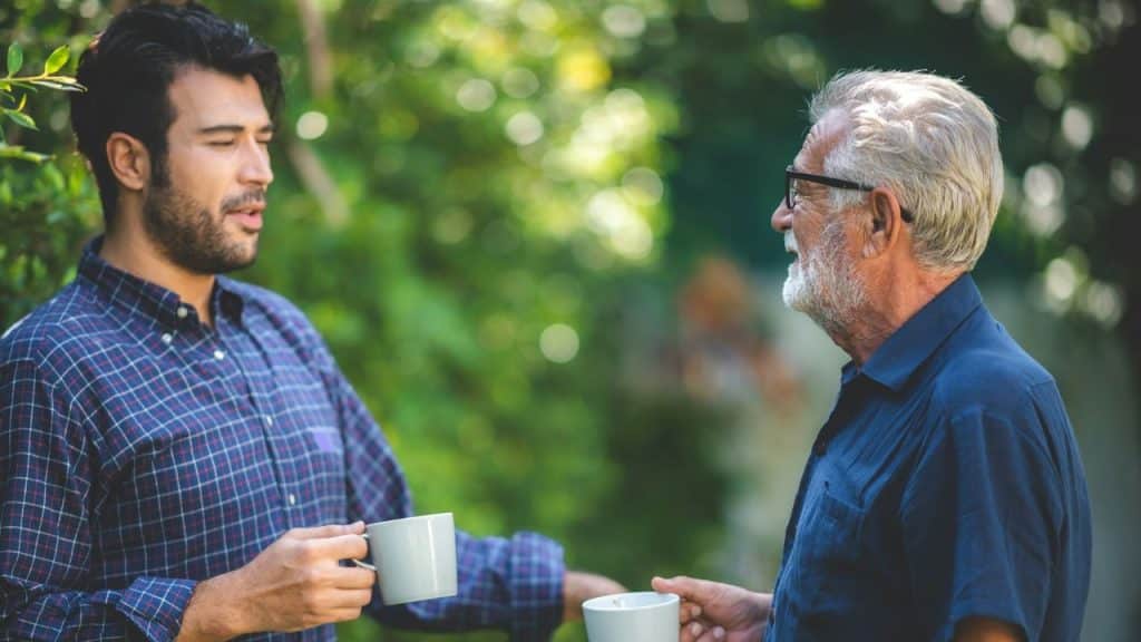 Two men stand outdoors holding mugs while facing each other and engaging in conversation.