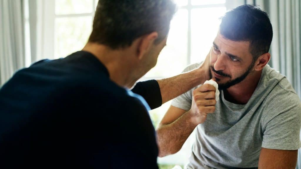 A man sits in front of another person who is holding a white tissue.