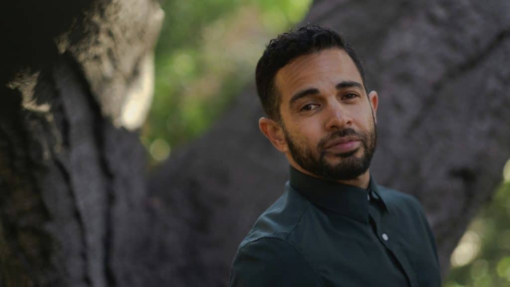 A man with dark hair and a beard looks toward the camera while standing outdoors.