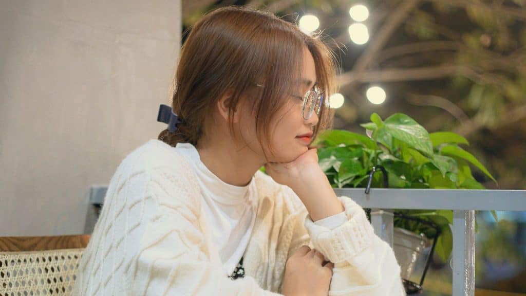 A woman wearing glasses and a white cardigan rests her chin on her hand.