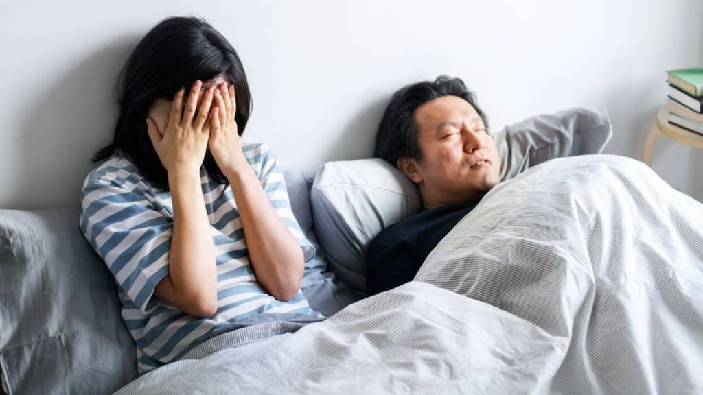 A woman sits in bed covering her face while a man sleeps soundly beside her.
