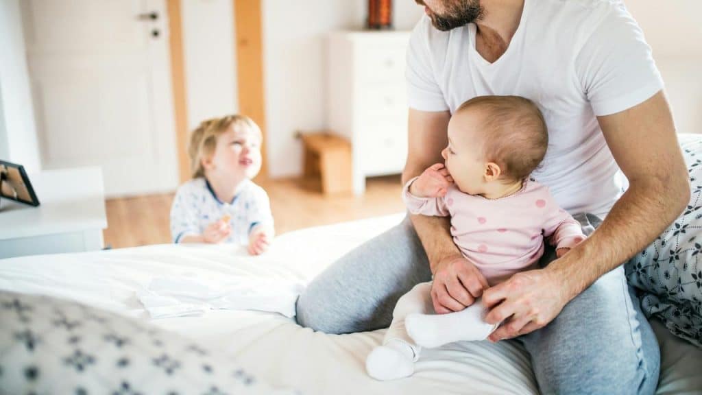 A man sitting on a bed holding a baby with a toddler nearby.