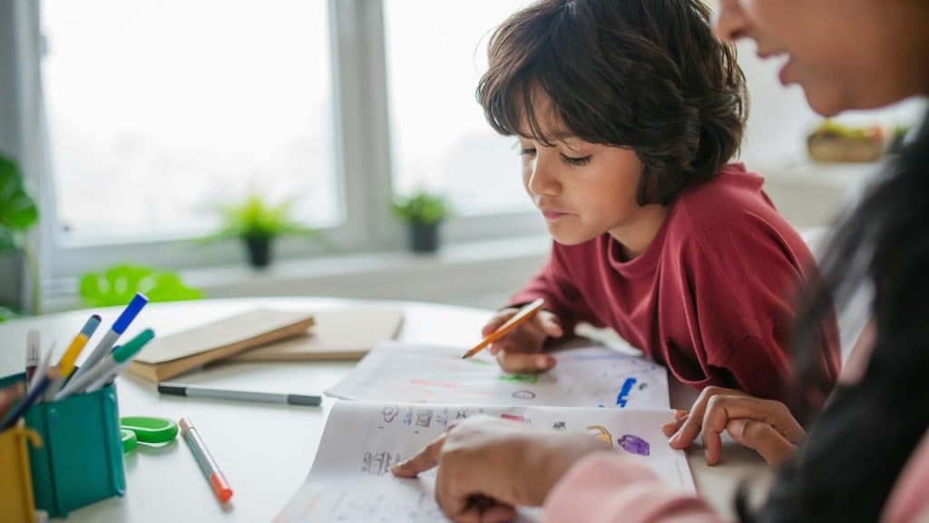 A child drawing in a notebook while an adult points to the page at a table.