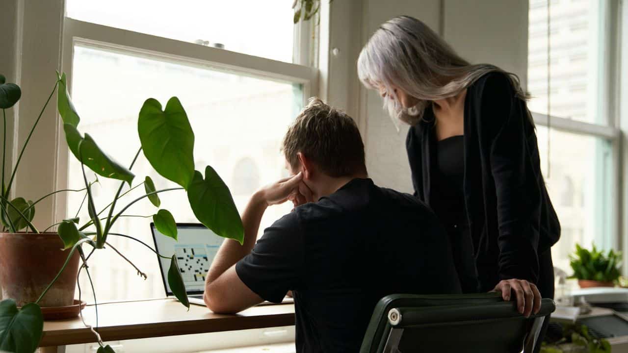 A man at a desk looks at a laptop while a woman stands beside him.
