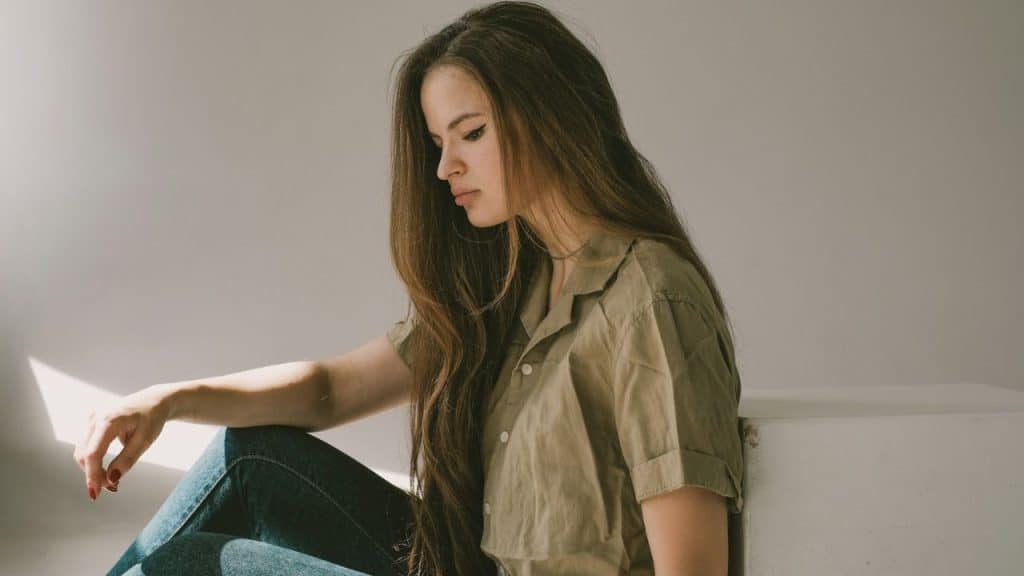 A young woman with long brown hair sits against a white wall, looking down with a sad expression.
