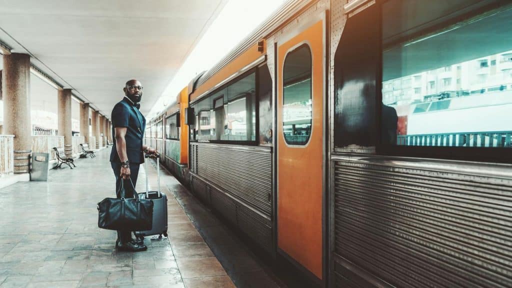A man in a suit standing on a train platform with luggage beside an orange train.