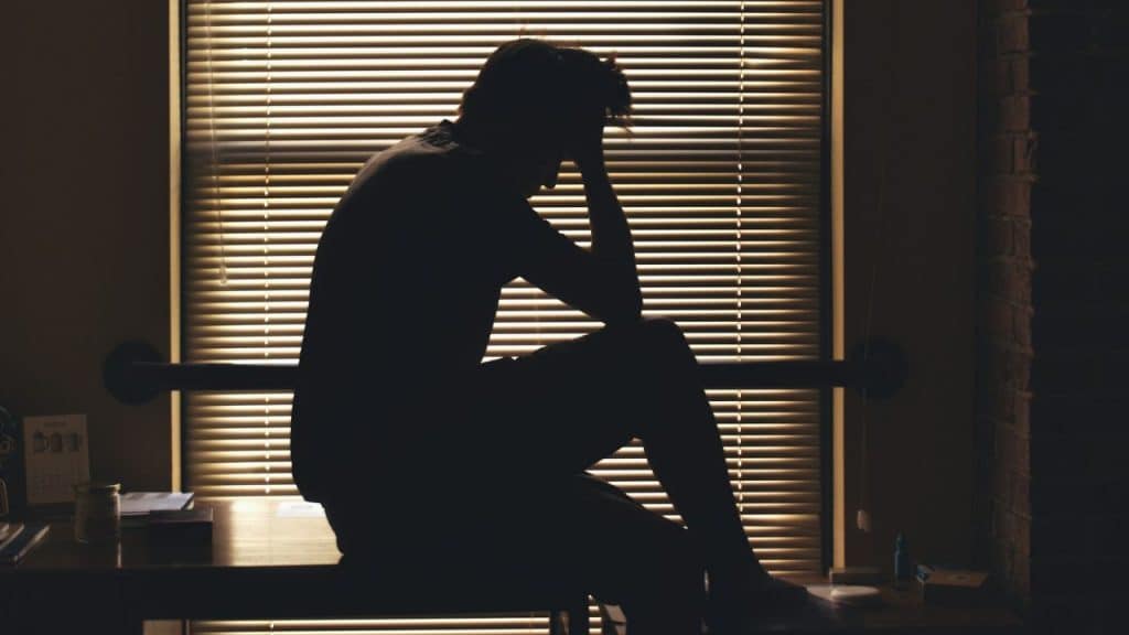 Silhouette of a person sitting on a desk, head in hands, against a window with blinds.